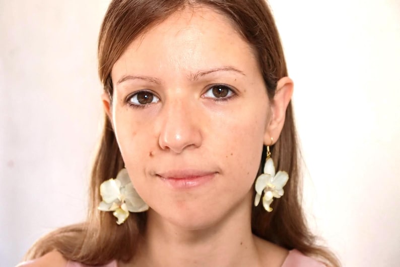 May include: A woman with long brown hair wearing white flower earrings. The earrings are made of white flowers with gold accents.