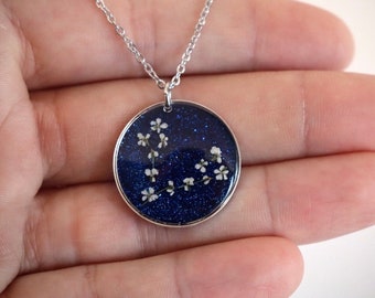 May include: A round silver pendant necklace featuring a deep blue glitter resin background embedded with small, delicate white pressed flowers. The circular charm hangs from a thin silver chain, creating a contrast between the dark, sparkling base and the light floral elements.