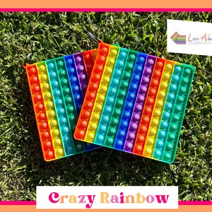 Puede incluir: Dos juguetes sensoriales pop-it de colores arcoíris con un lazo para llavero. Los juguetes están hechos de silicona y tienen un patrón de arcoíris con los números del 1 al 100 impresos en cada burbuja. Los juguetes están sobre una superficie de hierba verde. "Love Always Classroom" está impreso en una pequeña tarjeta en el fondo.