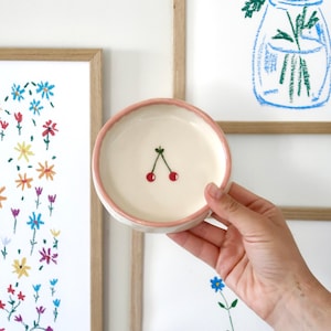 Puede incluir: Un plato pequeño y redondo de cerámica con interior crema y borde rosa. El plato presenta una sencilla ilustración de dos cerezas rojas con tallos verdes. El plato está sostenido en la mano de una persona, con obras de arte florales en el fondo.