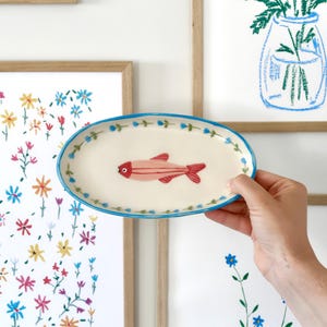 Könnte beinhalten: Ovale Keramikservierplatte mit handgemaltem roten Fischdesign. Die Platte hat eine cremefarbene Basis mit einem blauen Blumenrand. Der Fisch hat einen roten Körper und Schwanz mit einem einzelnen Auge. Die Platte wird vor gerahmter Blumenkunst gehalten.