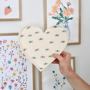 Puede incluir: Un plato de cerámica en forma de corazón, de color crema, con un patrón de pequeñas flores azules. El plato se sostiene frente a impresiones artísticas florales enmarcadas. Las impresiones presentan fresas, margaritas y otras flores silvestres.