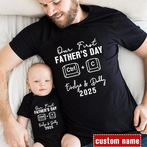 May include: A black t-shirt with white text that reads "Our First Father's Day Ctrl + C Evelyn & Daddy 2025". A baby is wearing a matching onesie with the text "Our First Father's Day Ctrl + V Evelyn & Daddy 2025".