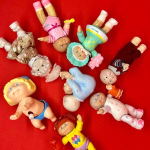 May include: A collection of small plastic dolls with various hair colors, facial expressions, and outfits, including dresses, sweaters, and pants. The dolls are arranged in a scattered pattern, featuring diverse accessories like hats and small toy figures.