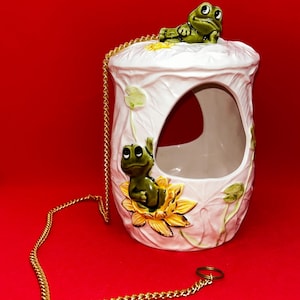Può includere: Un vaso sospeso in ceramica bianca con una catena dorata, con un design stravagante di due rane verdi. Una rana riposa in cima, l'altra è seduta su un fiore giallo. Il vaso ha una grande apertura e dettagli di foglie e fiori.