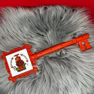 May include: An orange decorative key with a rectangular head featuring a colorful image of a coffee grinder. The key is lying on a gray, fluffy surface. The key's handle has a series of small holes.