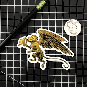 May include: A sticker featuring a whimsical winged monkey holding a golden chalice. The monkey has a smiling expression and is depicted in shades of brown and gold. The sticker is on a black grid background.