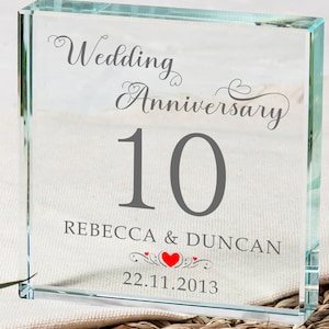 Può includere: Un blocco di vetro trasparente con il testo "Wedding Anniversary 10 Rebecca & Duncan 22.11.2013" inciso su di esso. Il numero 10 è in grigio e il resto del testo è in nero. Ci sono due cuori rossi con un design decorativo intorno a loro.