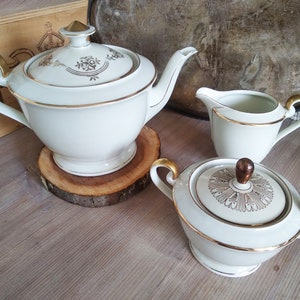 Puede incluir: Un juego de té de porcelana blanca con ribetes dorados. El juego incluye una tetera, una jarra de crema y un azucarero. La tetera tiene una tapa con un pomo dorado. La jarra de crema y el azucarero tienen detalles dorados en las asas.