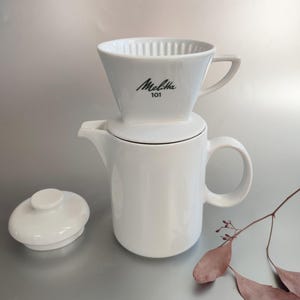 May include: A white ceramic pour-over coffee maker set. The set includes a cone-shaped filter holder with the text "Melitta 101", a matching teapot, and a lid. The coffee maker is designed for manual brewing.