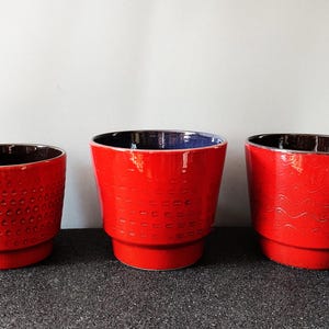 Könnte beinhalten: Drei rote Keramik-Blumentöpfe mit schwarzen Innenräumen. Die Töpfe haben unterschiedliche strukturierte Muster: eines mit kleinen Punkten, eines mit einem sich wiederholenden ovalen Design und eines mit wellenförmigen Linien. Die Töpfe stehen auf einer dunklen Oberfläche.