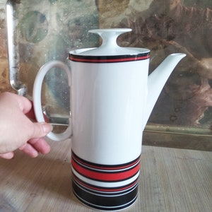 Puede incluir: Cafetera de cerámica blanca con un diseño de rayas negras y rojas. La cafetera tiene una tapa y un asa.