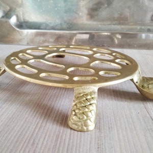 Puede incluir: Un posavasos de metal dorado en forma de tortuga con un diseño decorativo calado. El posavasos tiene cuatro patas y una cola.
