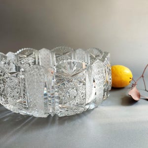 May include: A clear, heavy crystal bowl featuring intricate geometric cut patterns, including starburst motifs and cross-hatching. The bowl has a scalloped rim and a thick, faceted base, reflecting light through its detailed, pressed glass construction.