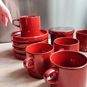 Puede incluir: Un juego de tazas y platillos de café de cerámica roja con una jarra de crema y un azucarero a juego. Las tazas y los platillos tienen un diseño simple y elegante.