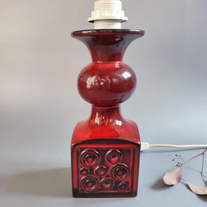 Könnte beinhalten: Ein glänzender roter Keramik-Lampenfuß mit einem quadratischen Unterteil, das geprägte kreisförmige Muster aufweist. Die Lampe hat einen bauchigen Mittelteil und eine trichterförmige Öffnung oben mit einer weißen Kunststoff-Lampenfassung.