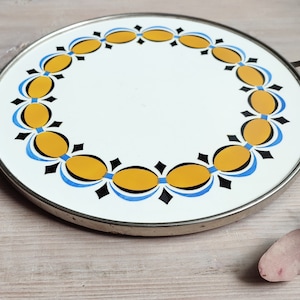 Pode incluir: Uma bandeja redonda de metal com uma superfície de esmalte branco e uma borda decorativa de ovais amarelos, azuis e pretos. A bandeja tem duas alças de metal.