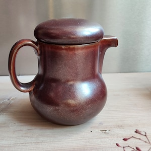 Puede incluir: Una jarra de crema de cerámica marrón con tapa. La jarra tiene un cuerpo redondeado y un asa. La tapa es ligeramente abovedada.