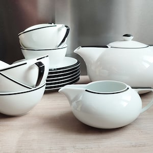 May include: A white teapot with a black trim and matching creamer and sugar bowl. The teapot has a round body and a spout on the side. The creamer and sugar bowl have a round body and a handle on the side. The set is made of porcelain and has a black trim around the rim.