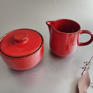 Könnte beinhalten: Eine rote Zuckerdose aus Keramik mit Deckel und ein passender Sahnekännchen. Die Zuckerdose ist rund mit Deckel und kleinem Griff. Der Sahnekännchen hat einen Griff und einen Ausguss. Beide Teile haben eine glänzende Oberfläche.