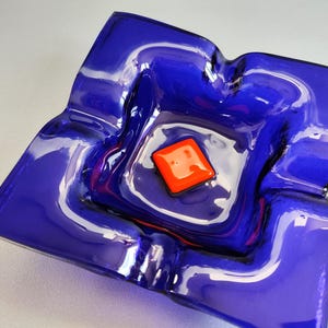 Könnte beinhalten: Quadratischer Aschenbecher aus kobaltblauem Glas mit glänzendem Finish. Der Aschenbecher hat ein zentrales, erhabenes Quadrat in leuchtendem Orange. Das Design ist eine moderne Interpretation eines klassischen Artikels.