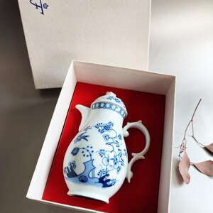 Puede incluir: Cafetera de porcelana blanca y azul con diseños florales y de pájaros, presentada en una caja blanca con interior rojo. La cafetera tiene un asa curvada y una tapa decorativa. La caja tiene un logotipo azul.