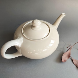 May include: A cream-colored ceramic teapot with a rounded body, a curved handle, and a spout. The lid has a small knob. The teapot is on a gray surface. A few dried leaves are in the background.