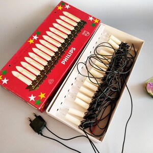 Peut inclure: Guirlande lumineuse de Noël vintage Philips en forme de bougies, dans une boîte rouge avec des étoiles blanches et des accents verts. La boîte est ouverte, révélant les lumières et le câblage noir. Une prise verte est visible.