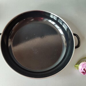 May include: A black enamel pan with two small handles. The pan has a silver rim and a dark interior. A pink flower with green leaves is in the lower right corner. The pan is likely used for cooking.