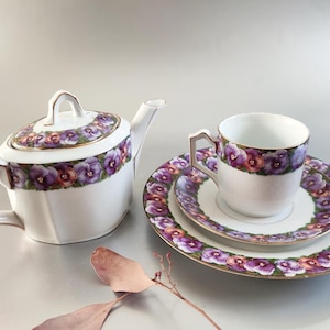Könnte beinhalten: Weißes Porzellan-Teeservice mit floralem Design aus lila und roten Stiefmütterchen. Das Set umfasst eine Teekanne, eine Tasse und zwei Teller. Die Ränder und Griffe sind goldfarben. Ein dekoratives Blatt liegt im Vordergrund.