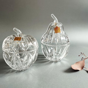 Könnte beinhalten: Zwei durchsichtige Glasbehälter mit Deckel in Form eines Apfels und einer Birne. Beide Stücke haben ein facettiertes, blattartiges Muster und einen kleinen Griff aus klarem Glas auf dem Deckel. Jeder Behälter ist mit einem kleinen, orangefarbenen Etikett versehen.