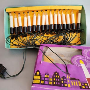 Peut inclure: Un ensemble de bougies de Noël vintage dans une boîte. Les bougies sont blanches avec des bases noires et des pointes transparentes. La boîte est turquoise et jaune, avec un couvercle de boîte violet représentant une silhouette de ville. Les lumières sont reliées par des fils verts.