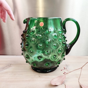 May include: A green glass jug with a textured surface. The jug has a handle and a round body.