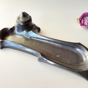 May include: A silver-coloured metal inkwell with a unique, abstract design. The inkwell has a raised, rounded section at one end and a long, shallow trough for ink.