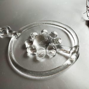 Könnte beinhalten: Klarer Glas-Servierteller mit einem kreisförmigen Griff und einem strukturierten zentralen Design. Der Teller hat einen ringförmigen Rand und einen dekorativen, abgerundeten Griff. Das Glas ist transparent und lässt Licht durch, auf einer silbernen Oberfläche.