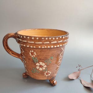 May include: Brown ceramic mug with a handle and three small feet. The mug features white dotted and dashed patterns around the rim and base, with floral designs in white and green. The interior is a light orange color.