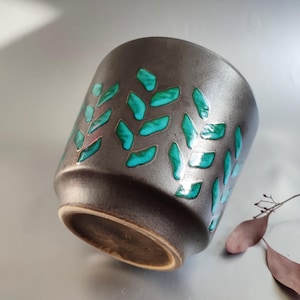 Peut inclure: Un pot de plante en céramique brun foncé avec un motif en forme de feuilles avec des accents turquoise et sarcelle. Le pot a une forme légèrement évasée et une surface texturée.