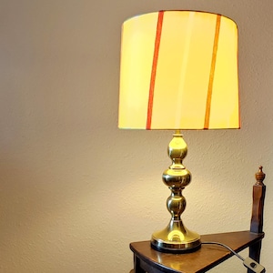 May include: A vintage brass table lamp with a yellow lampshade featuring vertical stripes in red and orange. The lamp has a bulbous base and is placed on a small wooden table. The lamp is plugged in and ready to use.