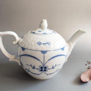 Puede incluir: Tetera de porcelana blanca con diseños florales y geométricos azules. La tetera tiene un asa curva, un pico y una tapa con un pequeño adorno en forma de pájaro. El diseño incluye elementos florales en relieve.