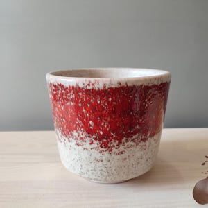 Può includere: Una tazza in ceramica con un esterno rosso e bianco strutturato. La tazza ha una forma cilindrica ampia con una base bianca macchiata che si trasforma in una vivace fascia rossa. Il bordo è anch'esso bianco.