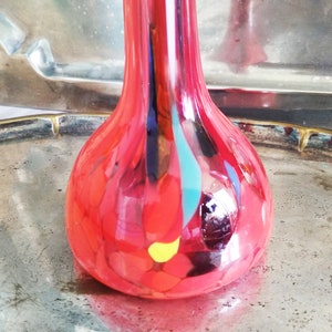 Peut inclure: Un vase en verre rouge avec un motif en spirale de bleu, jaune et noir. Le vase a un corps rond et un col étroit.