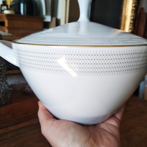Puede incluir: Tazón de servir de porcelana blanca con un borde dorado y un patrón geométrico gris. El tazón tiene dos asas y una tapa.