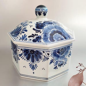 Può includere: Una scatola ottagonale in ceramica bianca con un coperchio, decorata con motivi floreali blu e bianchi di Delft. Il coperchio ha un pomo in cima.