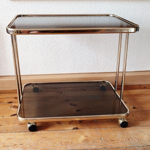 Könnte beinhalten: Ein goldfarbener Servierwagen aus Metall mit zwei Glasböden. Der Wagen hat vier Räder und steht auf einem Holzboden.