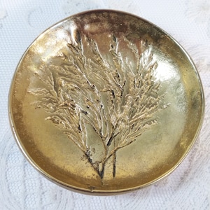 Könnte beinhalten: Kleine, runde, goldfarbene Schale mit einem erhabenen, strukturierten Design eines Pflanzen- oder Astzweigs in der Mitte. Die Schale hat einen leicht gebogenen Rand und eine glatte, reflektierende Oberfläche.