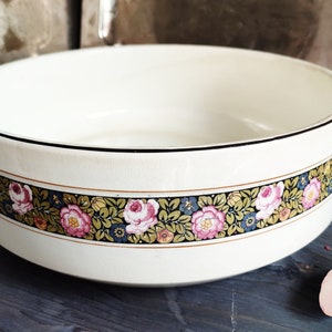 Könnte beinhalten: Eine weiße Keramikschüssel mit schwarzem Rand und einem Blumendesign mit rosa Rosen, grünen Blättern und goldenen Akzenten. Die Schüssel steht auf einer dunkelblauen Holzoberfläche.