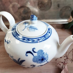 Könnte beinhalten: Weiße Porzellan-Teekanne mit blauen Blumen- und Blättermustern. Die Teekanne hat einen runden Körper, einen gebogenen Ausguss und einen Schlaufengriff. Der Deckel ist mit einem blauen Blumenknauf versehen.