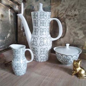 Könnte beinhalten: Ein Set aus drei weißen Keramik-Küchenutensilien mit einem grauen Blumenmuster. Das Set beinhaltet eine Milchkännchen, eine Zuckerdose mit Deckel und eine Kaffeekanne.