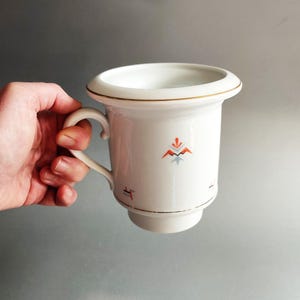 May include: A white ceramic mug with a wide, flared rim and a curved handle. The mug features a delicate gold trim along the rim and base, accented by small, geometric patterns in orange, black, and light blue printed on the exterior surface.