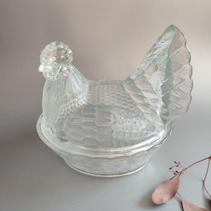 May include: A clear glass hen-shaped dish with a textured surface. The dish is designed with intricate details, including a comb, feathers, and a round base. The lid of the dish is shaped like the hen's body, and the base is a shallow bowl.
